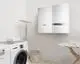 Wandmontierter Warmwasserspeicher in Weiß von Vaillant, ausgestattet mit einem Bedienfeld. Im Hintergrund befinden sich eine Waschmaschine und ein Wäschekorb.